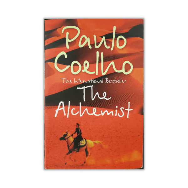 The Alchemist by Paulo Coelho - The Paperhound Online Bookshop. Secondhand books for sale online in South Africa. A copy of 'The Alchemist' by Paulo Coelho featuring a distinctive cover design. The background showcases a desert landscape bathed in warm tones of orange and red, evoking a sense of adventure and mysticism. In the foreground, a lone figure rides a horse across the sandy expanse, symbolizing the journey theme of the novel. The author's name, 'Paulo Coelho,' is prominently displayed in large, white script at the top, with the subtitle 'The International Bestseller' in smaller lettering beneath it. The title 'The Alchemist' appears in a similar white script below the author's name. - The Paperhound Online Bookshop. Secondhand books for sale online in South Africa.