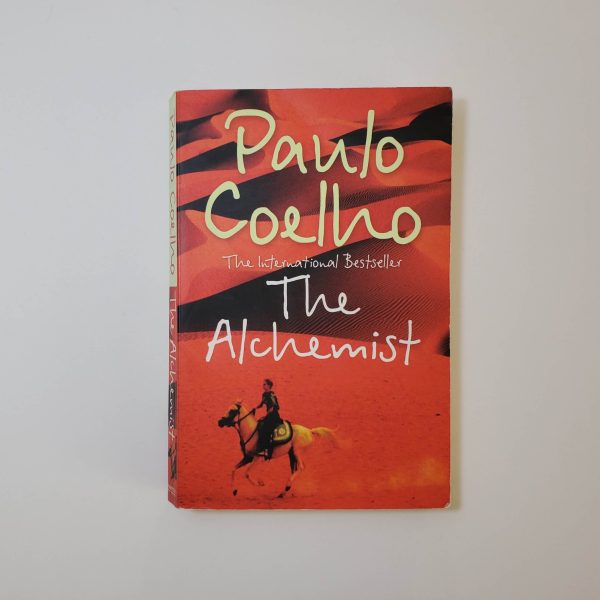 The Alchemist by Paulo Coelho - The Paperhound Online Bookshop. Secondhand books for sale online in South Africa. A copy of 'The Alchemist' by Paulo Coelho featuring a distinctive cover design. The background showcases a desert landscape bathed in warm tones of orange and red, evoking a sense of adventure and mysticism. In the foreground, a lone figure rides a horse across the sandy expanse, symbolizing the journey theme of the novel. The author's name, 'Paulo Coelho,' is prominently displayed in large, white script at the top, with the subtitle 'The International Bestseller' in smaller lettering beneath it. The title 'The Alchemist' appears in a similar white script below the author's name. - The Paperhound Online Bookshop. Secondhand books for sale online in South Africa.